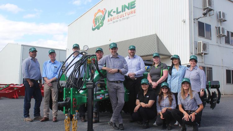 Twelve staff members standing in front of a K-Line Ag machine in the carpark at the k-Line Cowra factory. The K-Line Ag name and logo is printed on the front of the building.